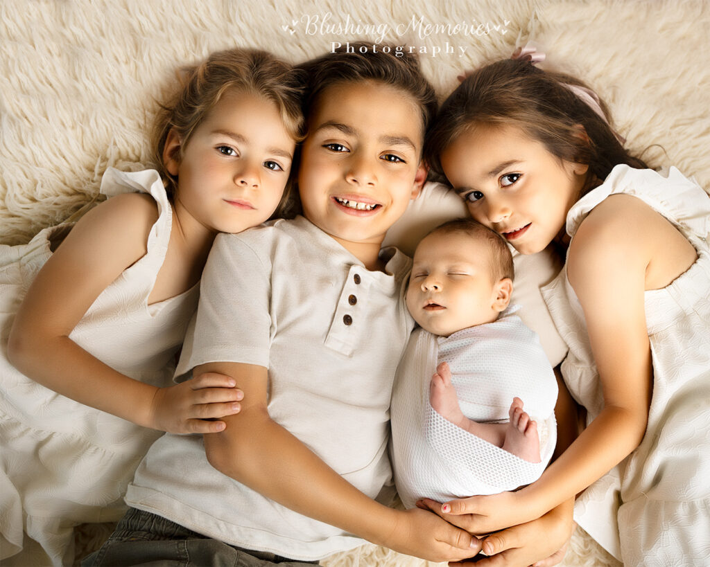 Three siblings cuddling their newborn baby brother wrapped in a white swaddle on a soft cream flokati rug, captured at Blushing Memories Photography in El Dorado Hills
