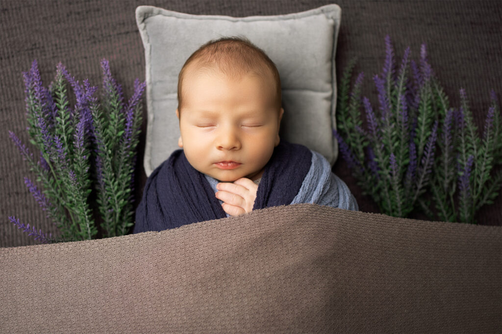 A newborn baby boy peacefully sleeping, wrapped in a soft blue wrap with delicate flowers beside him, photographed at Blushing Memories Photography Studio in El Dorado Hills, California.