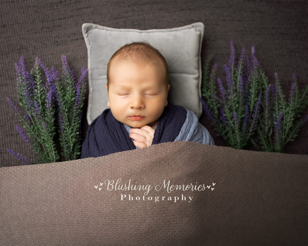 A newborn baby boy peacefully sleeping, wrapped in a soft blue wrap with delicate flowers beside him, photographed at Blushing Memories Photography Studio in El Dorado Hills, California.