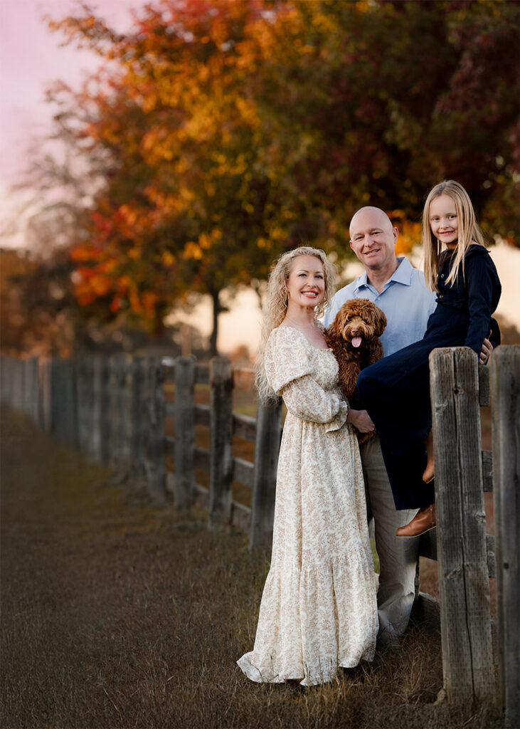 Outdoor fall family portrait beside a wooden fence at sunset, with parents, daughter sitting on the fence, and their dog – Blushing Memories Photography, El Dorado Hills, Folsom, Sacramento, Bay Area