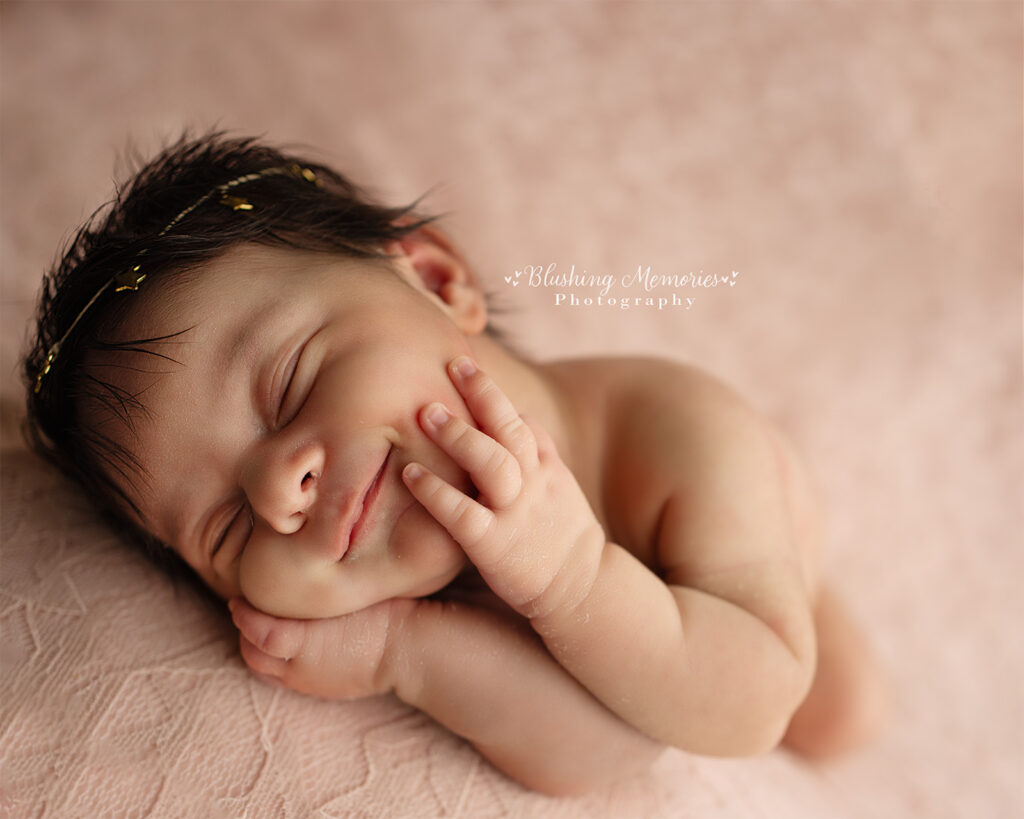 Newborn baby girl sleeping peacefully with hands on cheeks on a soft pink blanket, professional newborn photography by Blushing Memories Studio in Folsom California.
