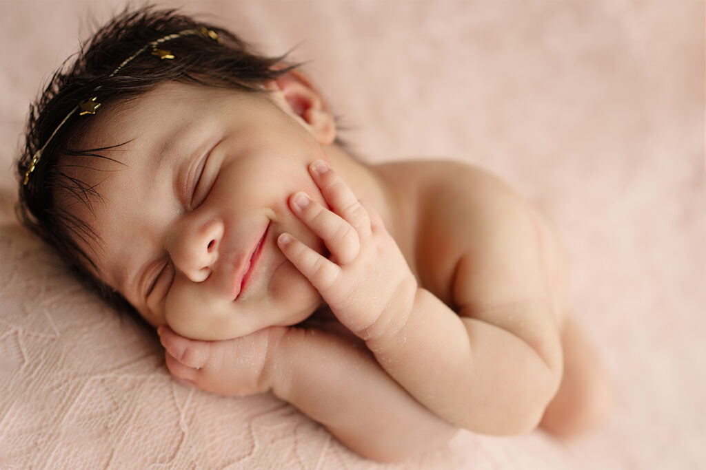 Newborn baby girl sleeping peacefully with hands on cheeks on a soft pink blanket, professional newborn photography by Blushing Memories Studio in Folsom California.