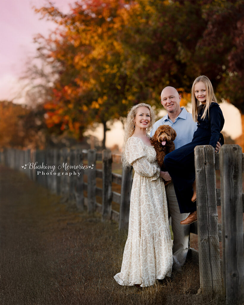 Outdoor fall family portrait beside a wooden fence at sunset, with parents, daughter sitting on the fence, and their dog – Blushing Memories Photography, El Dorado Hills, Folsom, Sacramento, Bay Area