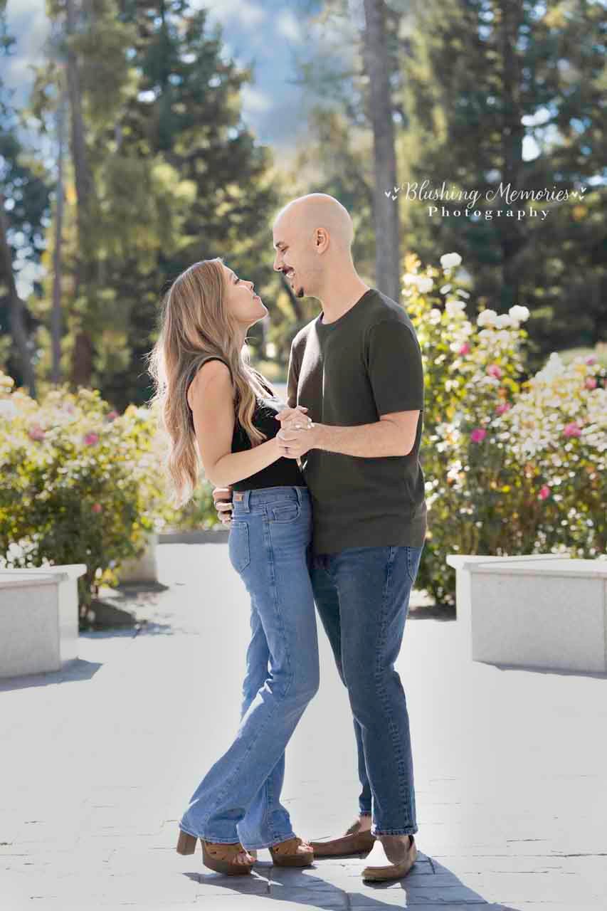 A couple in casual clothing holds hands and gazes at each other in a sunny garden setting, captured by Blushing Memories Photography.