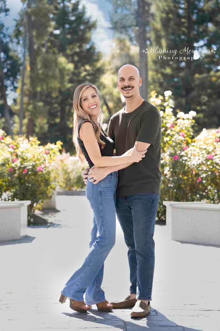 A smiling couple posing together in a sunny garden, wearing casual outfits of jeans and t-shirts, captured by Blushing Memories Photography.