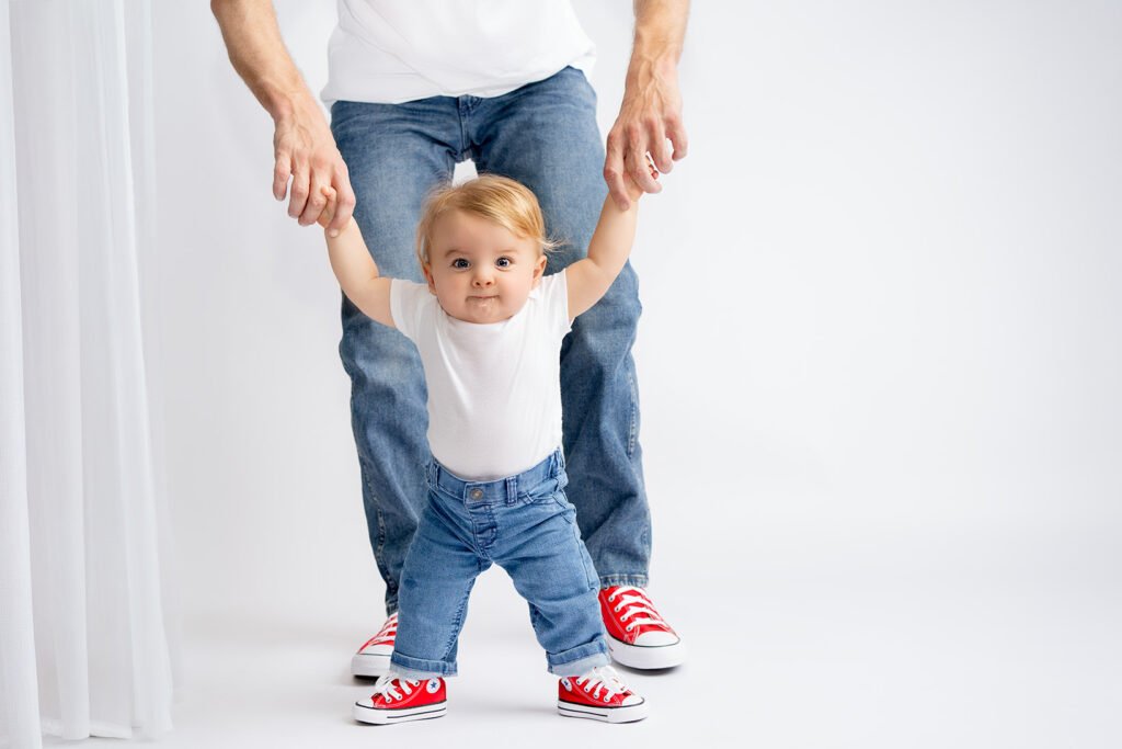 A father from Elk Grove holds his baby's hands during a milestone session at El Dorado Hills Blushing Memories photo studio, both wearing jeans and red sneakers.