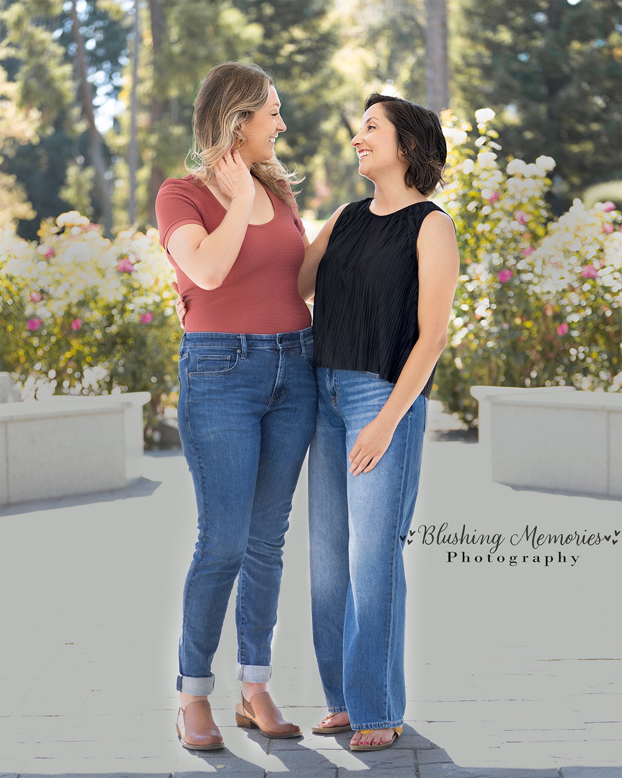 Two women smiling at each other during a couple photo session in a sunny Roseville garden with blooming flowers in the background.