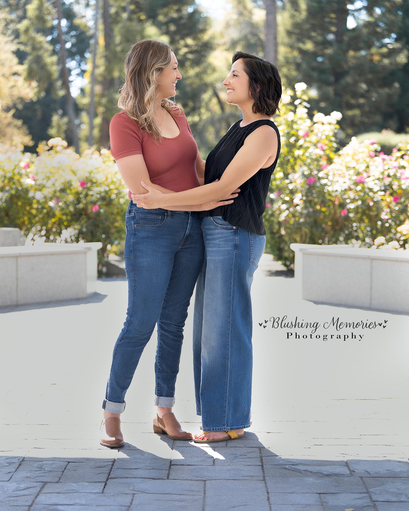 Two women smiling and embracing during a couple photo session in a sunny Roseville garden, wearing casual tops and blue jeans.