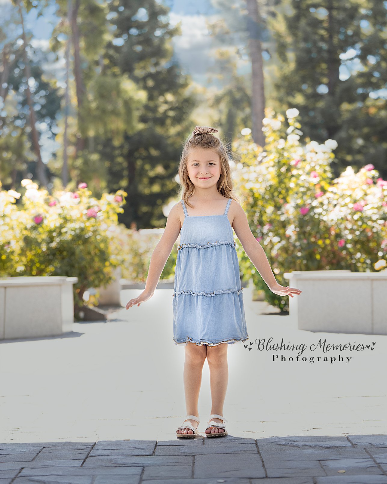 A young girl in a blue denim dress poses outdoors for a Blushing Memories studio girl photo session in a bright, sunlit Roseville garden setting.