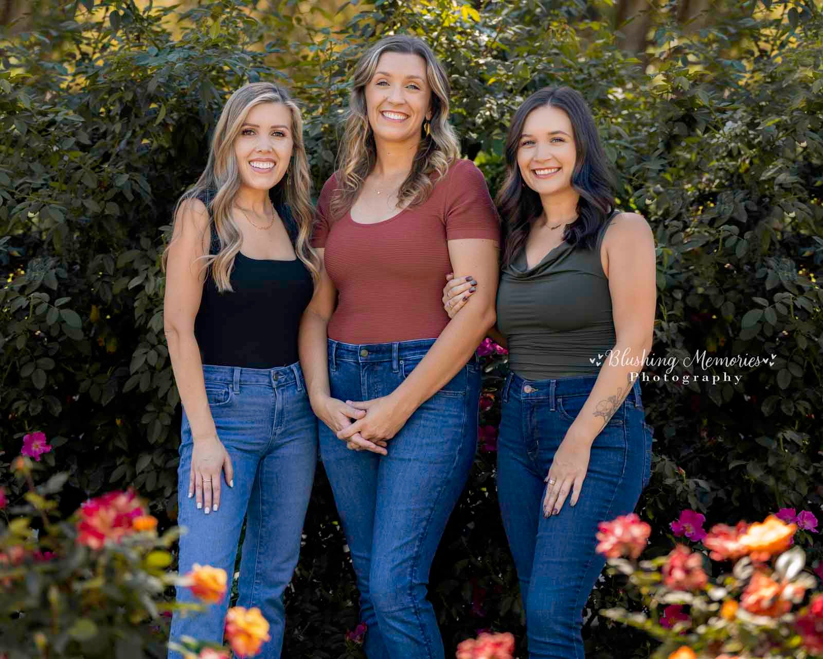 Three smiling sisters from Roseville pose together during an outdoor photo session in Sacramento, standing in front of a lush green rose bush.