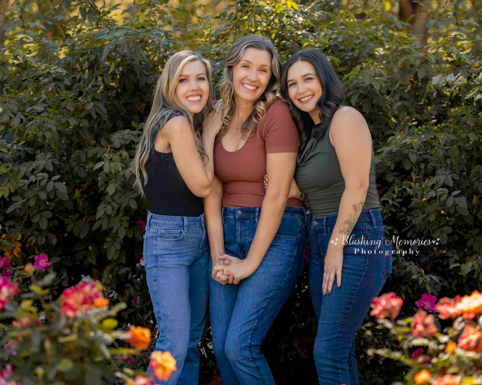 Three smiling sisters from Roseville pose together during an outdoor photo session in Sacramento, wearing casual tops and blue jeans in a garden.